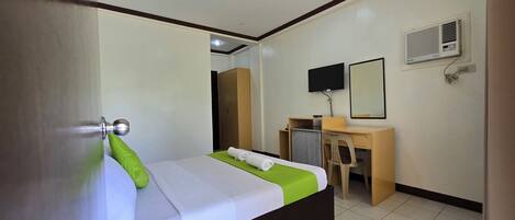 A hotel room with a bed, a desk, a chair, a TV, and an air conditioning unit.