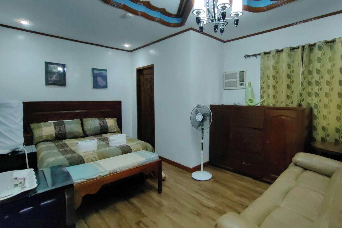 A bedroom with a bed, a wooden headboard, a bedside table, a fan, a sofa, and a window with curtains.