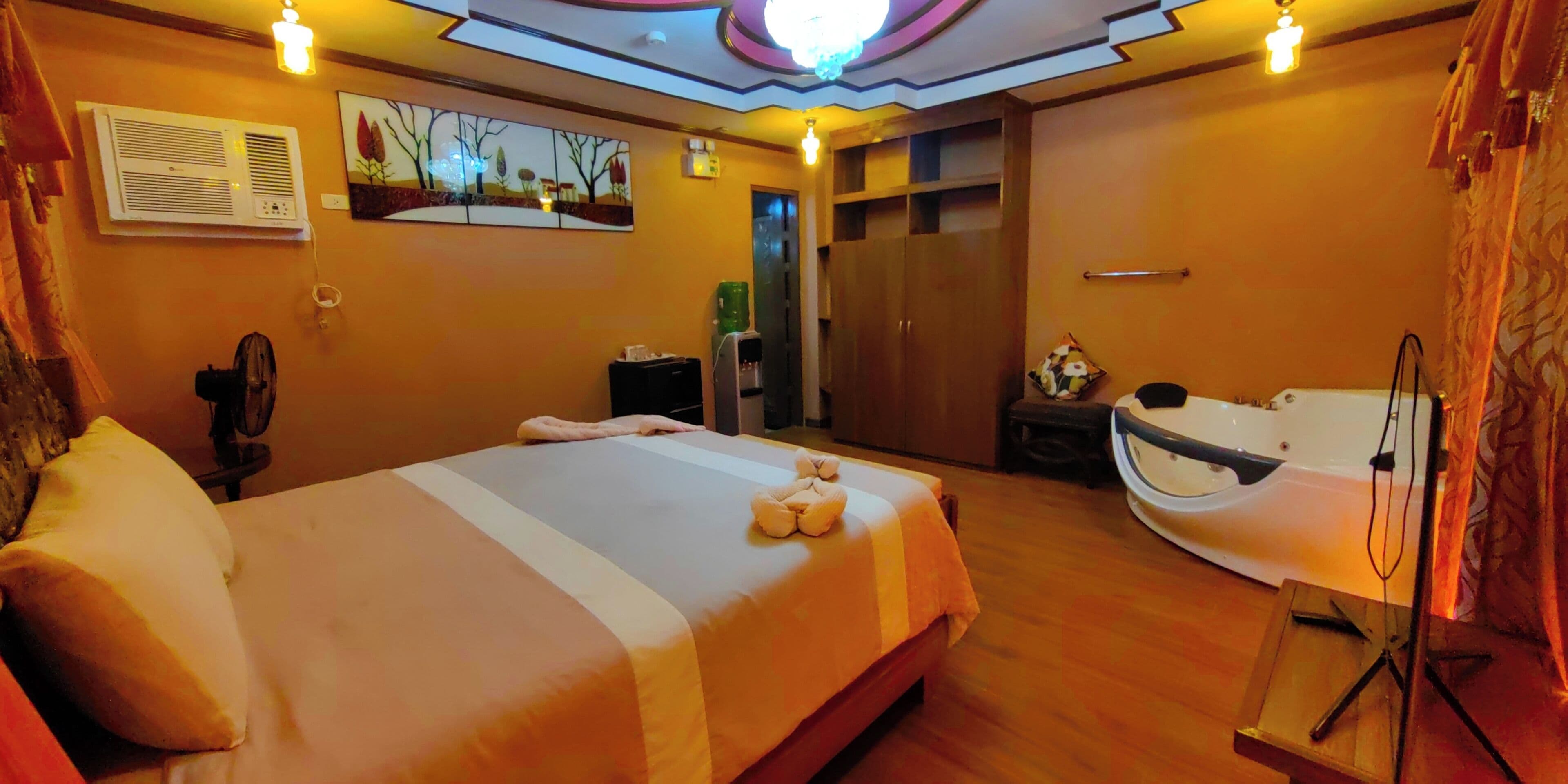 A hotel room with a large bed, a wooden floor, a wall-mounted air conditioner, a painting, a wooden wardrobe, and a bathtub.