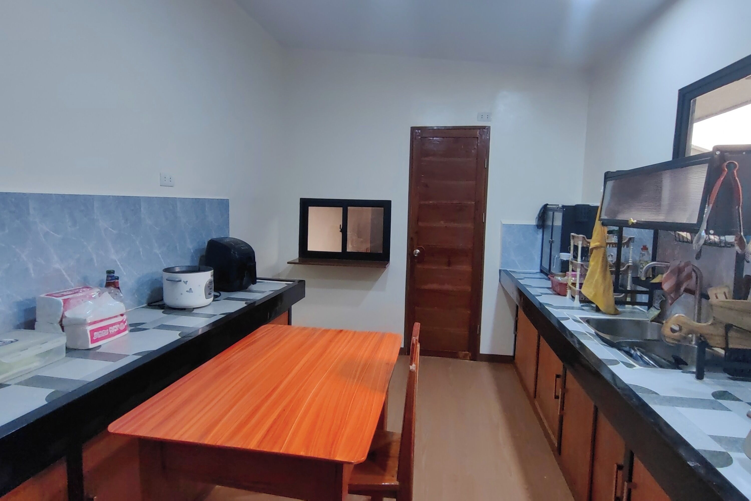 Una cocina funcional con una mesa de madera y un banco, un microondas, una tetera y un lavabo con un paño amarillo.