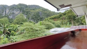 A view from a covered area with a red railing, overlooking a lush green landscape with dense vegetation and a prominent cliff in the background. - Summer Wind Pension House El Nido (El Nido)