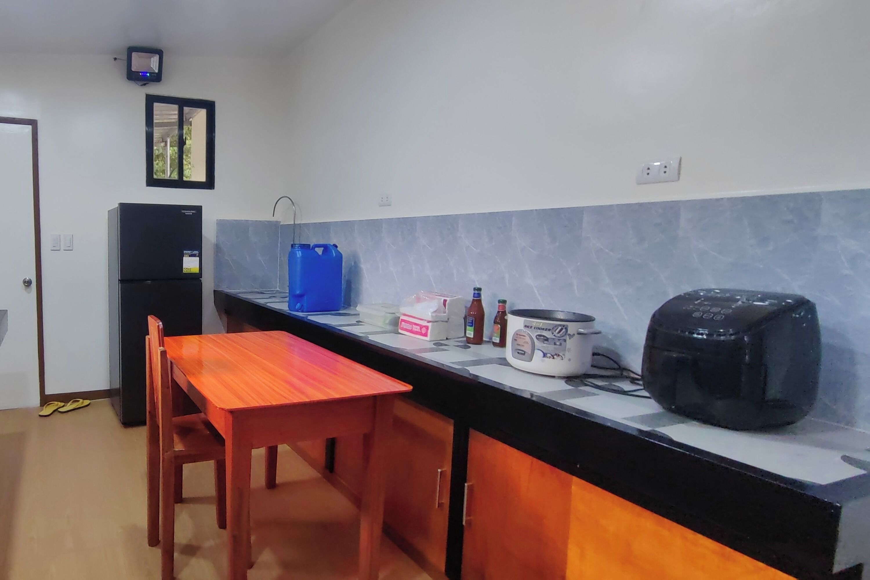A compact kitchen with a refrigerator, a blue water dispenser, a rice cooker, and a toaster.
