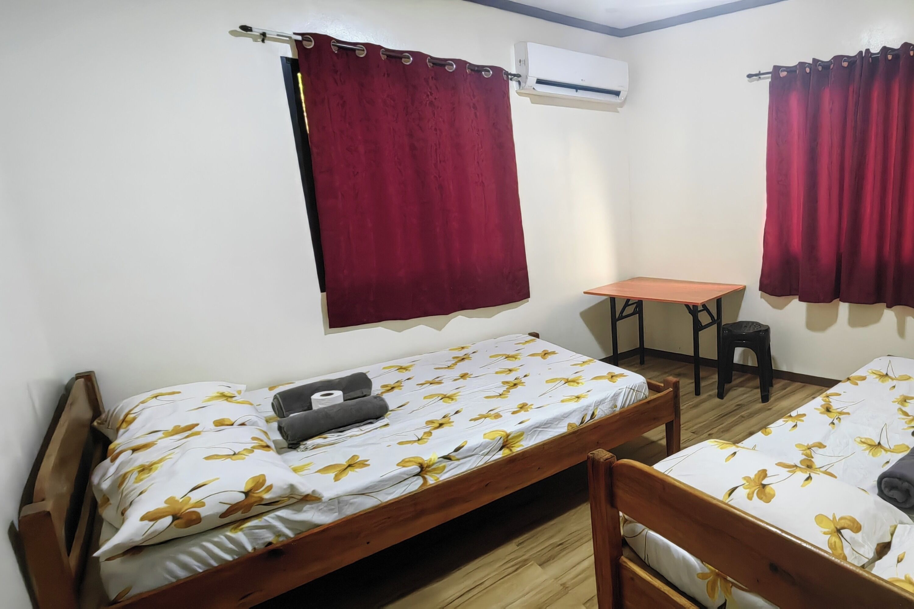 A room with two wooden beds, a small table, a chair, and an air conditioning unit on the wall.