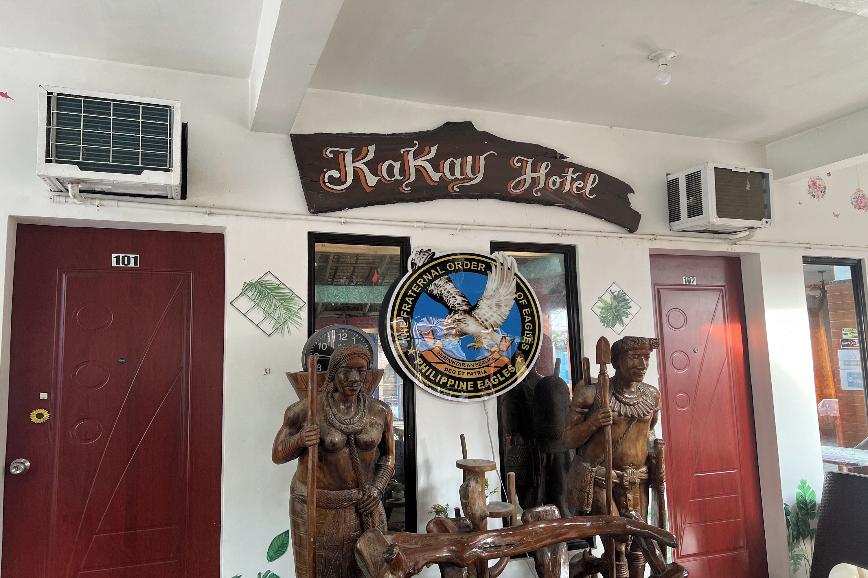 Entrada de un hotel con un cartel que dice "Kakay Hotel" y un logo del águila filipina. Hay dos puertas de madera, una con la etiqueta "101" y la otra con "107".