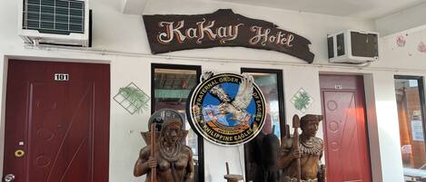 Entrada de un hotel con un cartel que dice "Kakay Hotel" y un logo del águila filipina. Hay dos puertas de madera, una con la etiqueta "101" y la otra con "107".