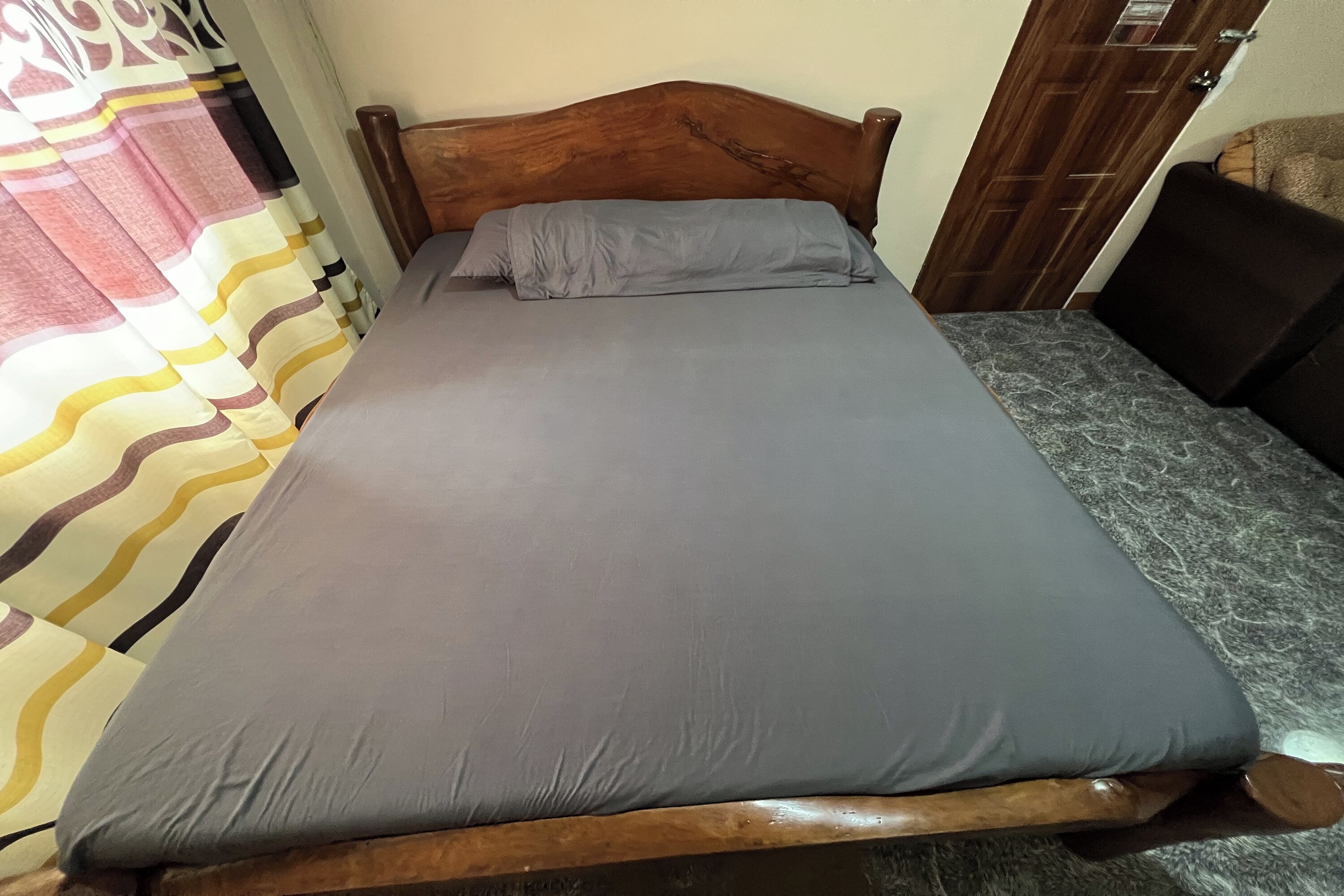 Una cama con cabecera de madera, ropa de cama gris y cortina estampada.