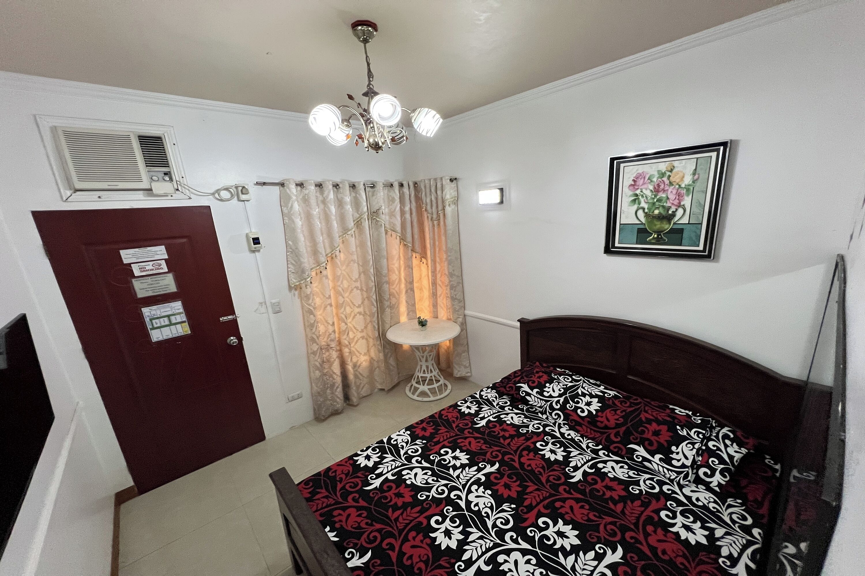 Un dormitorio con una cama, una mesita de noche, un espejo, una puerta, una ventana con cortinas y un equipo de aire acondicionado instalado en la pared.