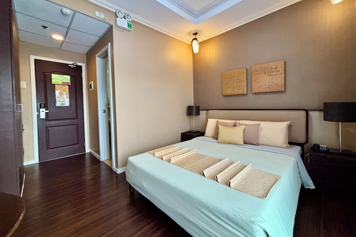 A hotel room with a large bed, a nightstand, a lamp, and a door leading to another room.