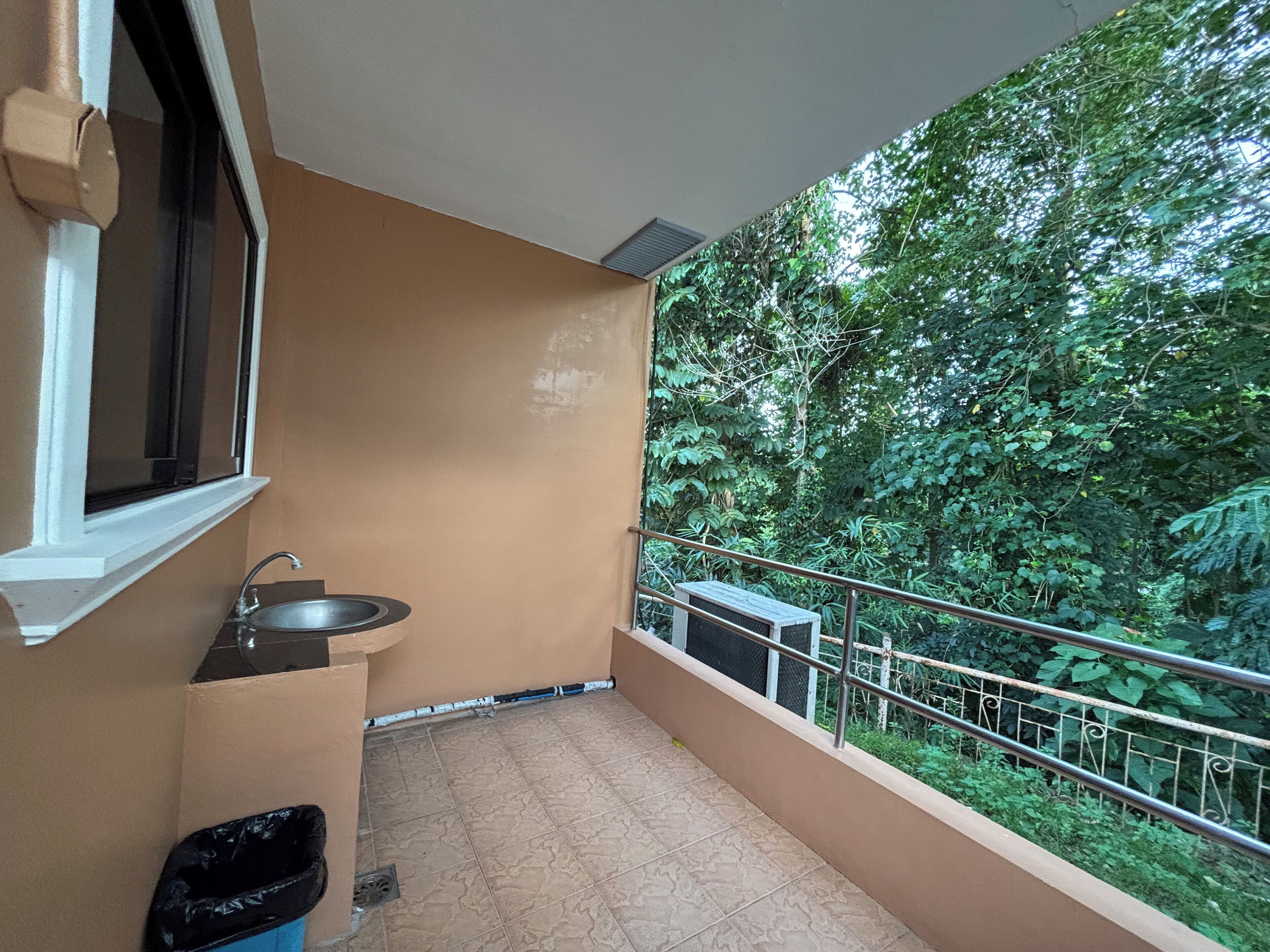 A balcony with a sink and a trash bin, surrounded by dense greenery.