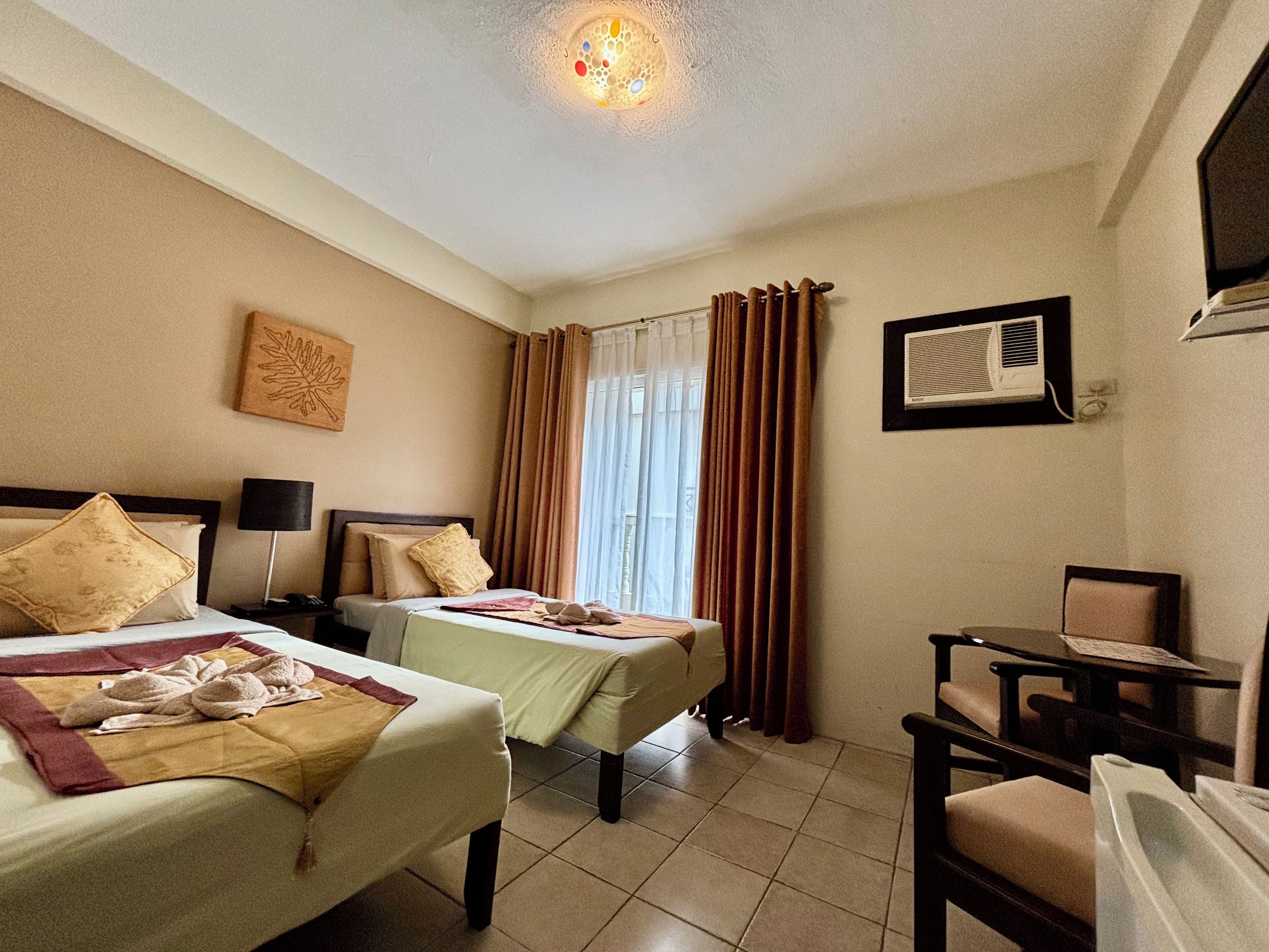 A hotel room with two beds, a desk, a chair, a ceiling light, and a window with curtains.