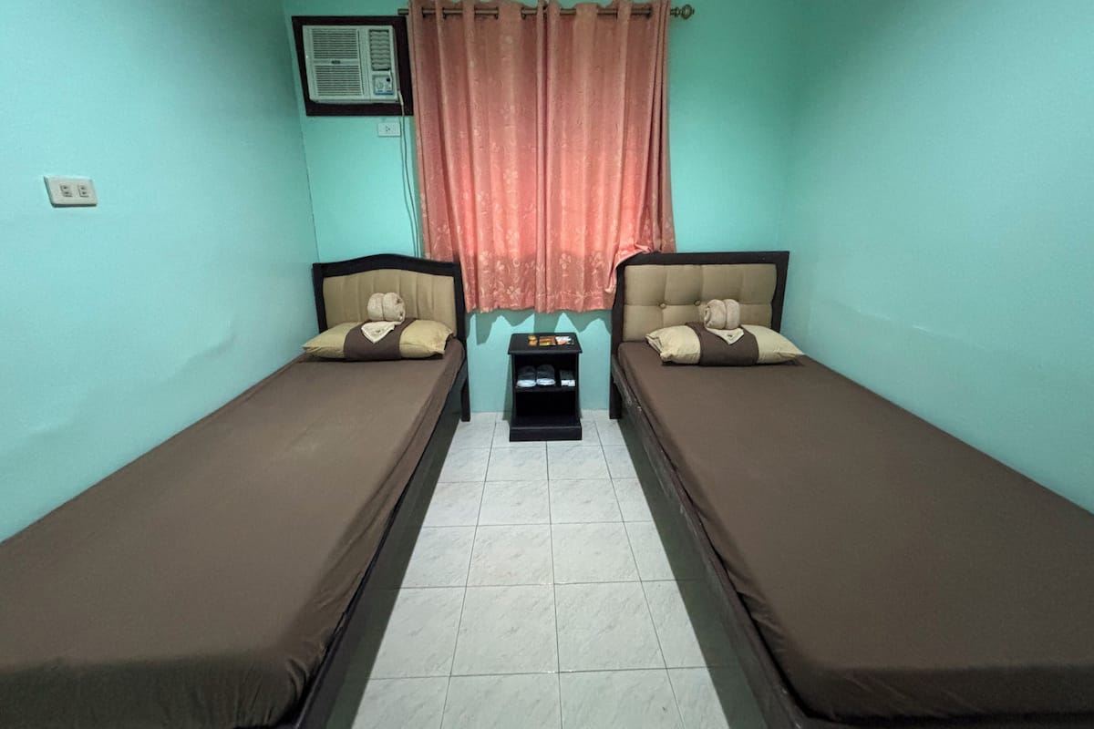 A twin room with two single beds, a small table, a shelf with items, and a window with curtains.