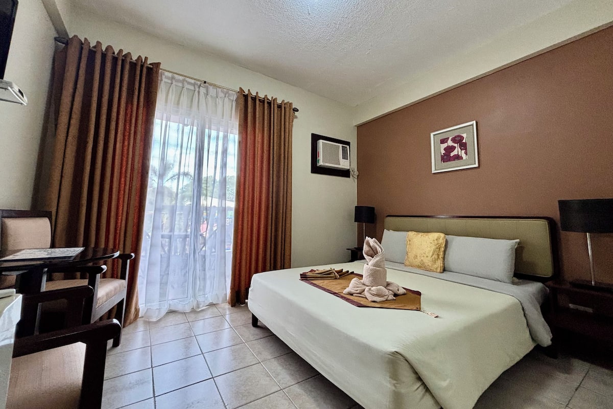 A hotel room with a bed, a desk, and a chair. There is a window with curtains, a wall-mounted air conditioner, and a framed picture on the wall.