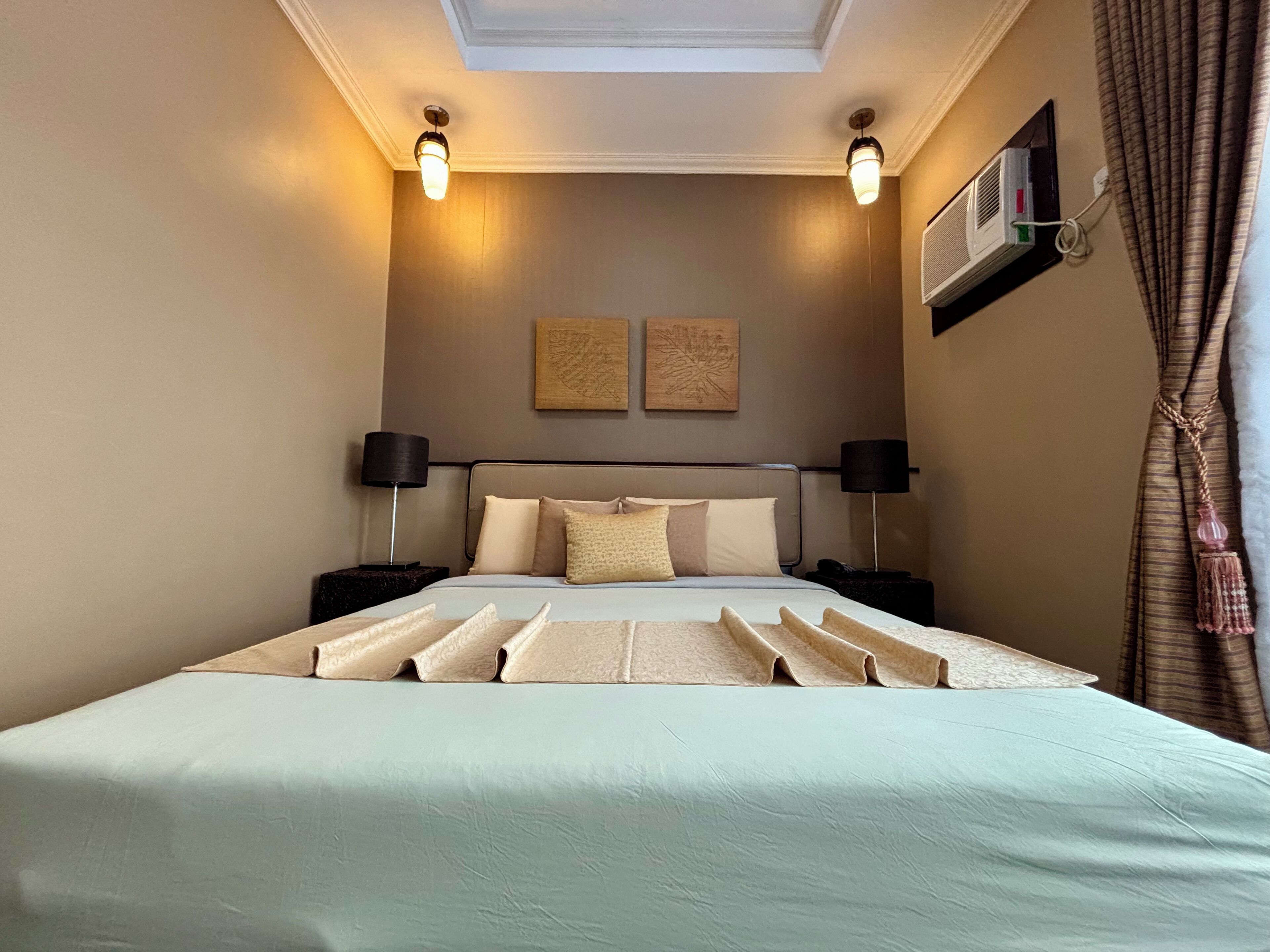 Ein Hotelzimmer mit einem großen Bett, zwei Nachttischen, einer Deckenleuchte, einer Klimaanlage und einem fenster mit Vorhängen.