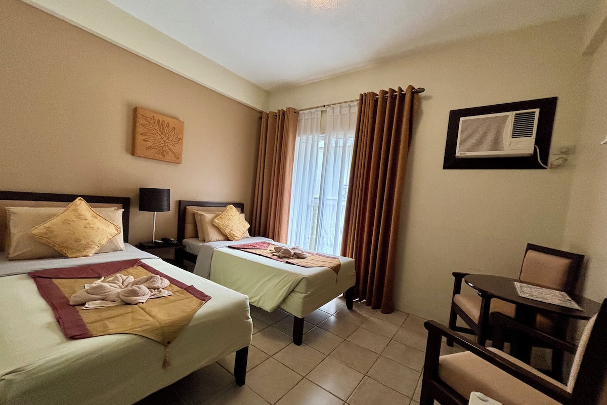 A hotel room with two beds, a desk, a chair, an air conditioning unit, and a window with curtains.