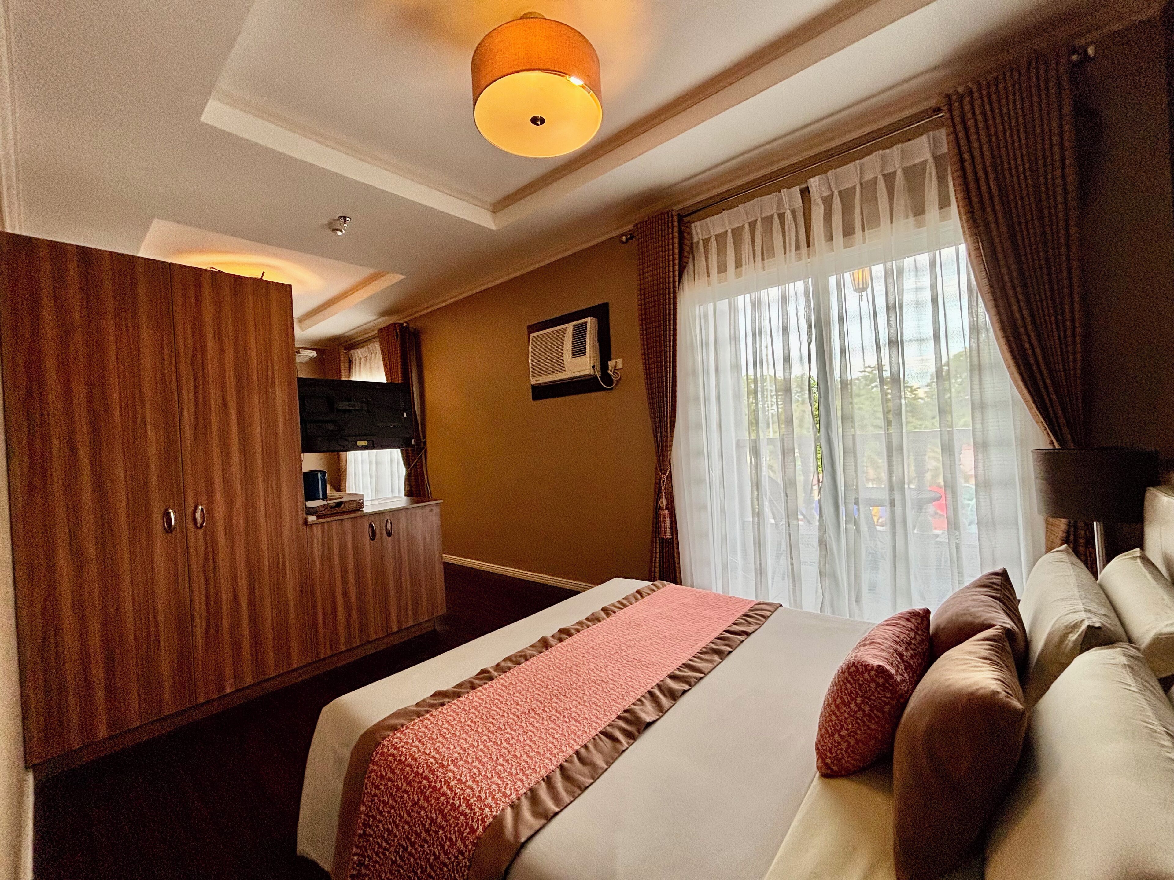 A hotel room with a bed, a wooden wardrobe, a television, and a window with curtains.