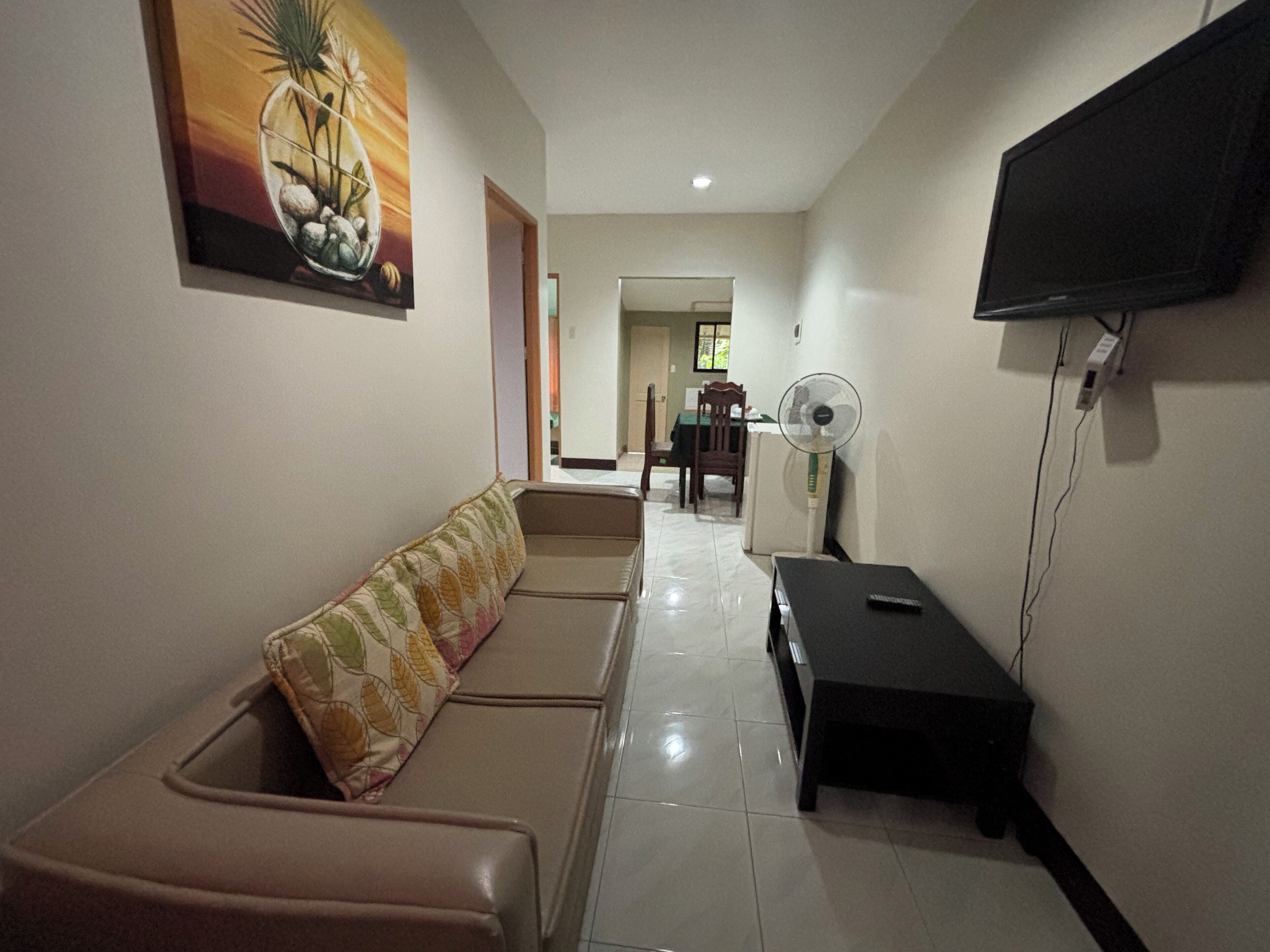 A living room with a sofa, a flat-screen TV mounted on the wall, a fan, and a painting on the wall.