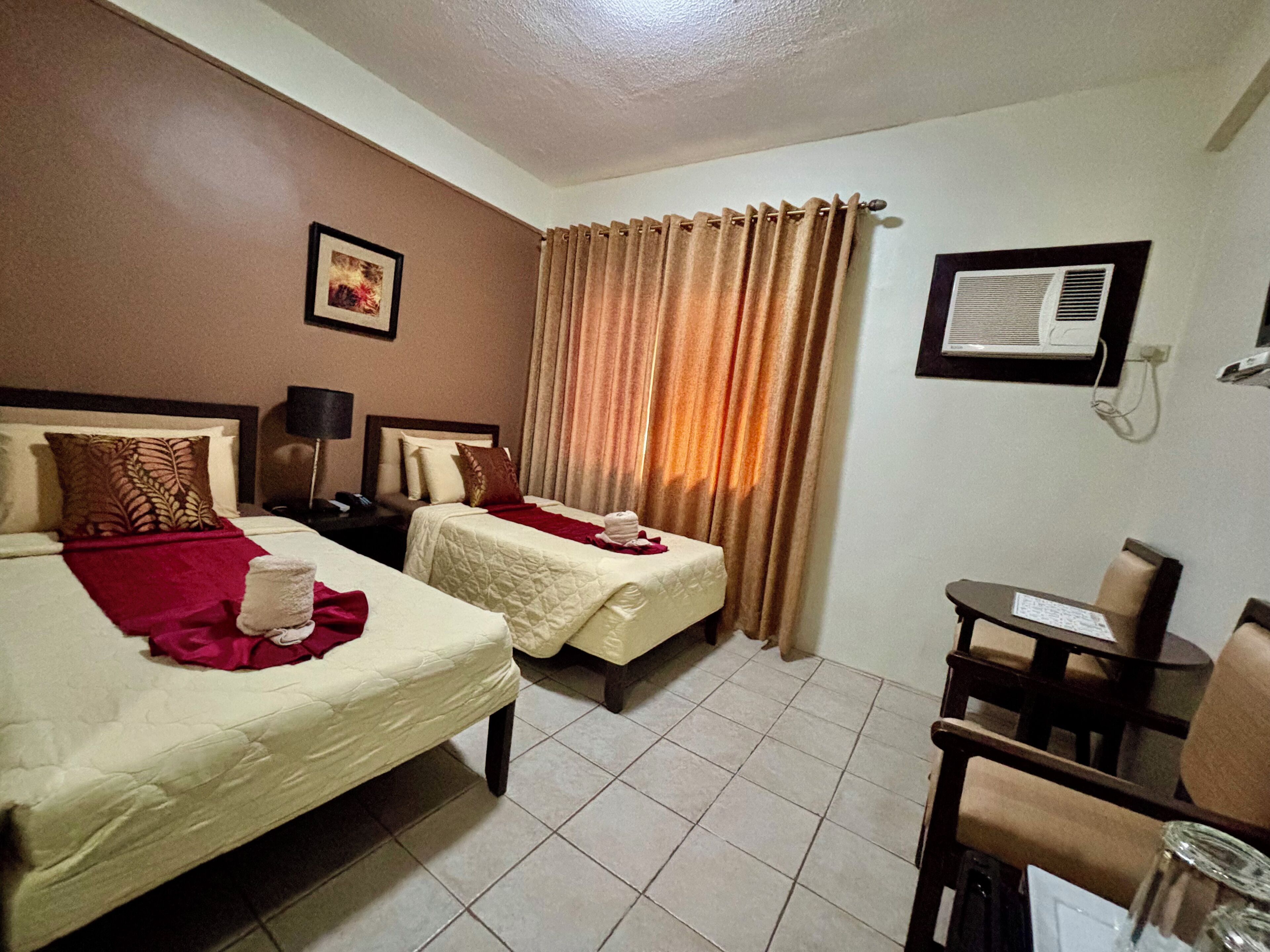 Habitación de hotel con dos camas, un escritorio, una silla, un equipo de aire acondicionado y una cortina.