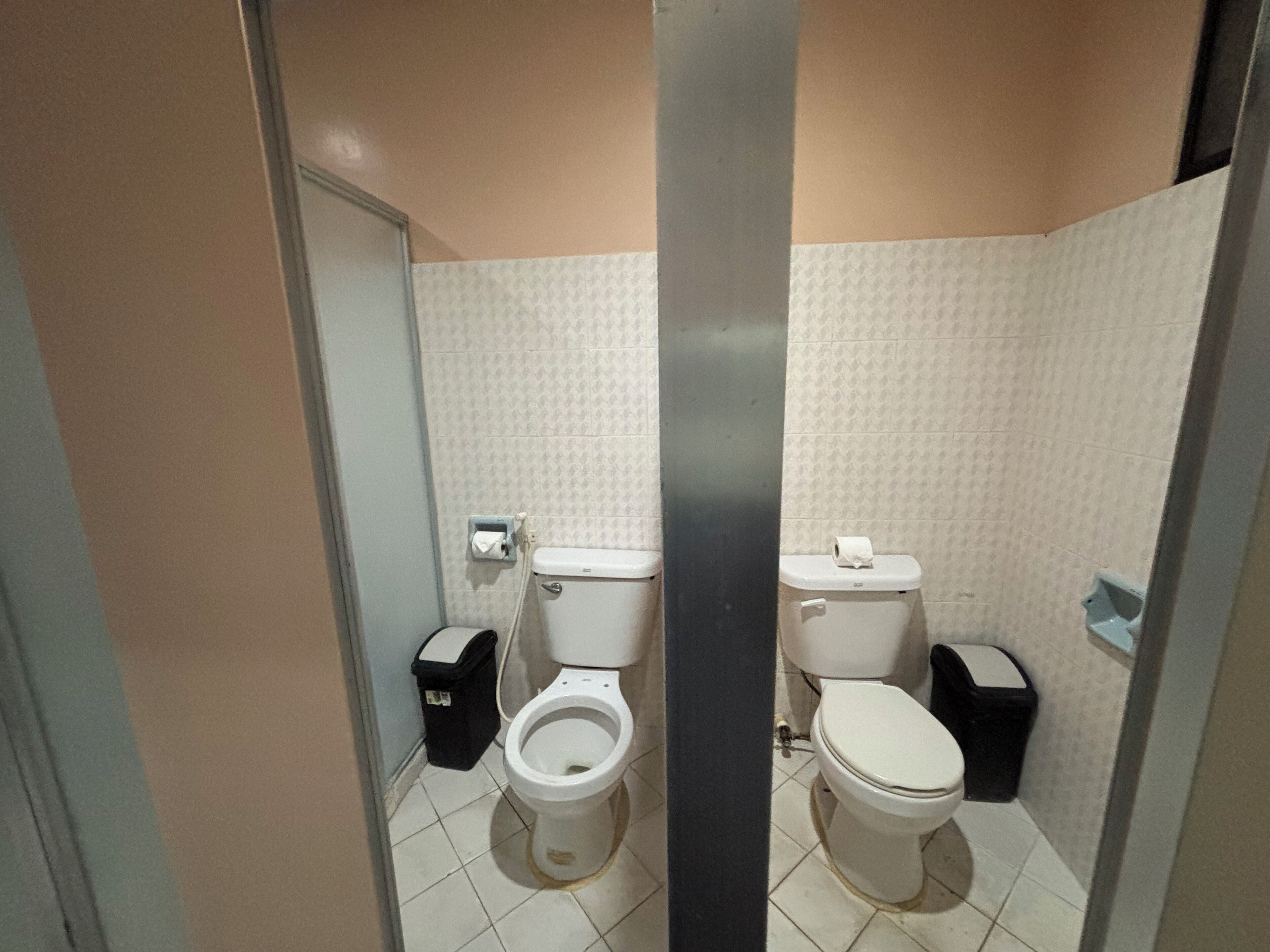 A small, tiled bathroom with two toilets, a trash bin, and a paper towel dispenser.