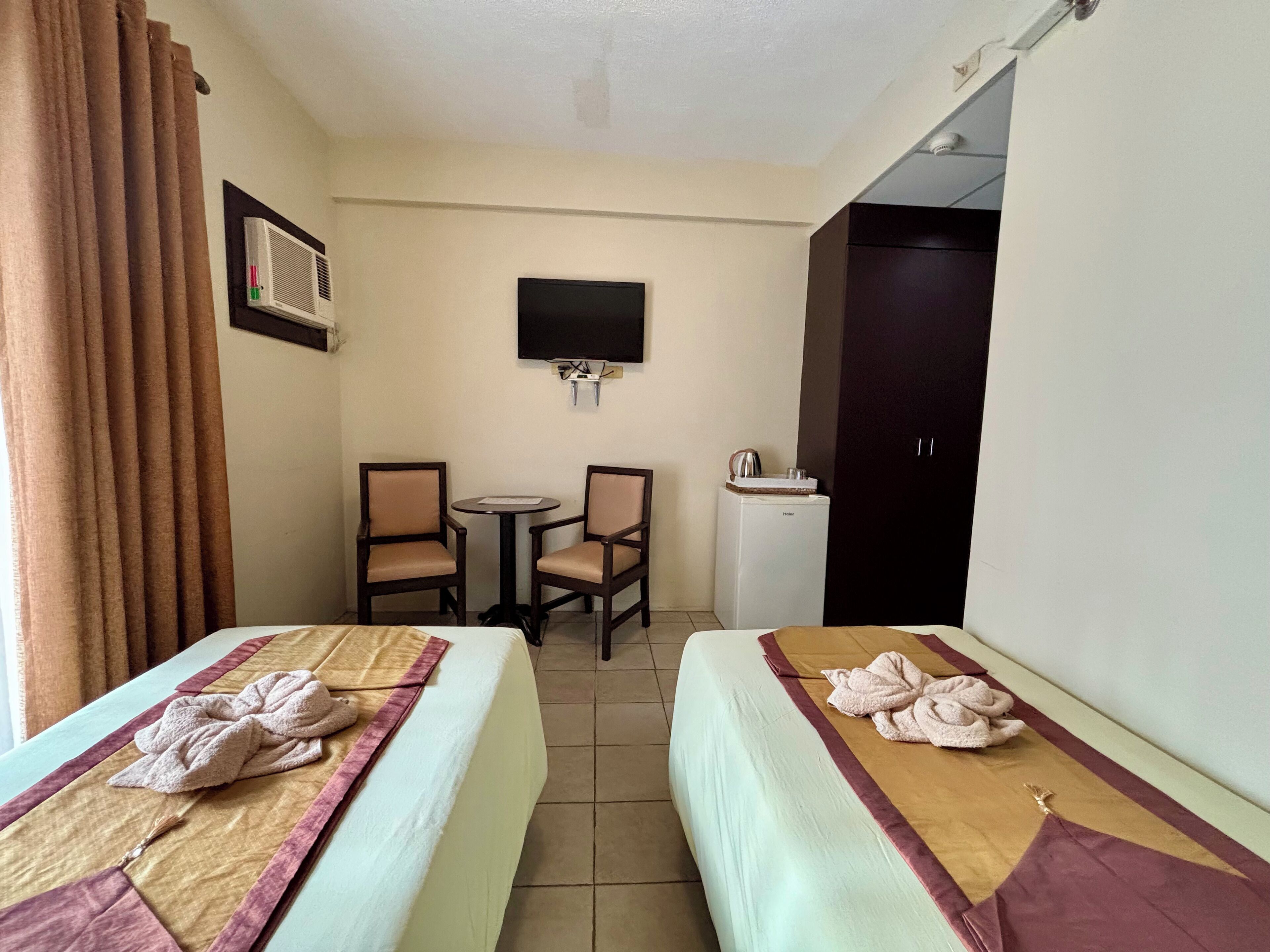 Habitación de hotel con dos camas, una mesita con sillas, un televisor montado en la pared y un refrigerador.