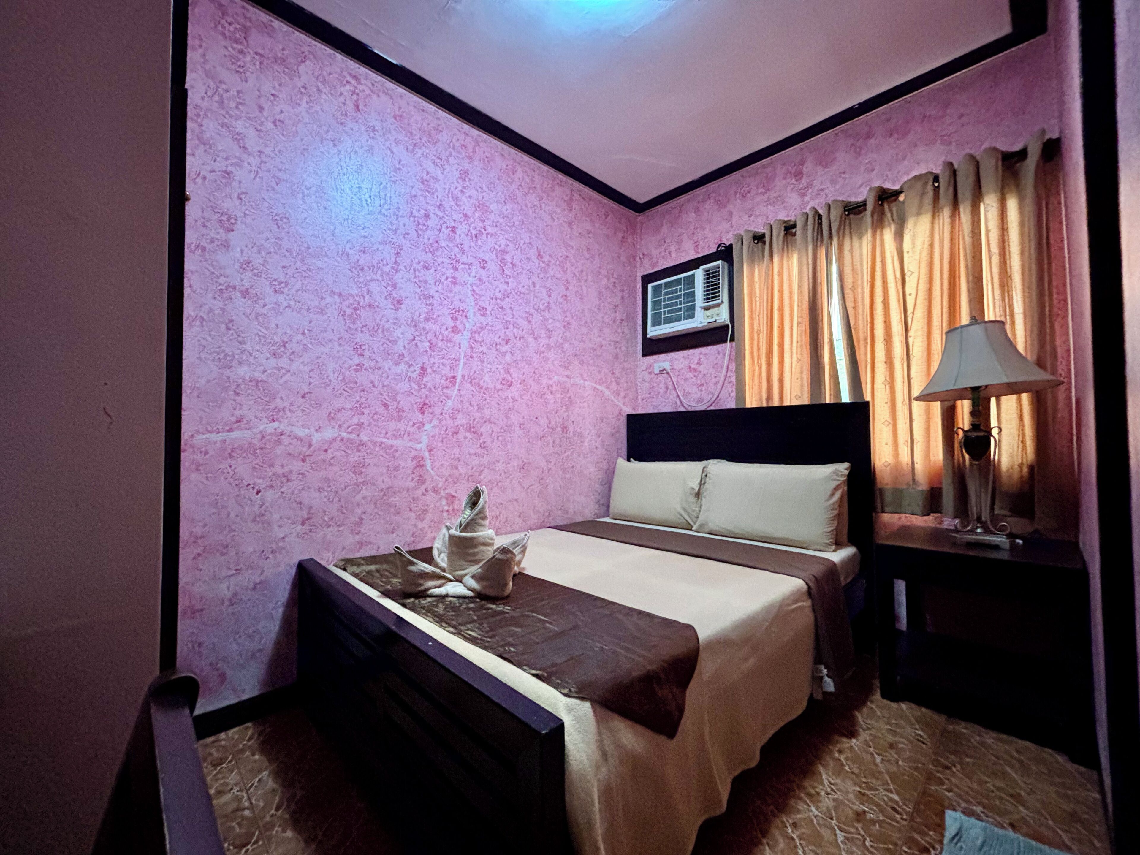 A room with pink walls, a bed with a headboard, a bedside table with a lamp, and an air conditioning unit.