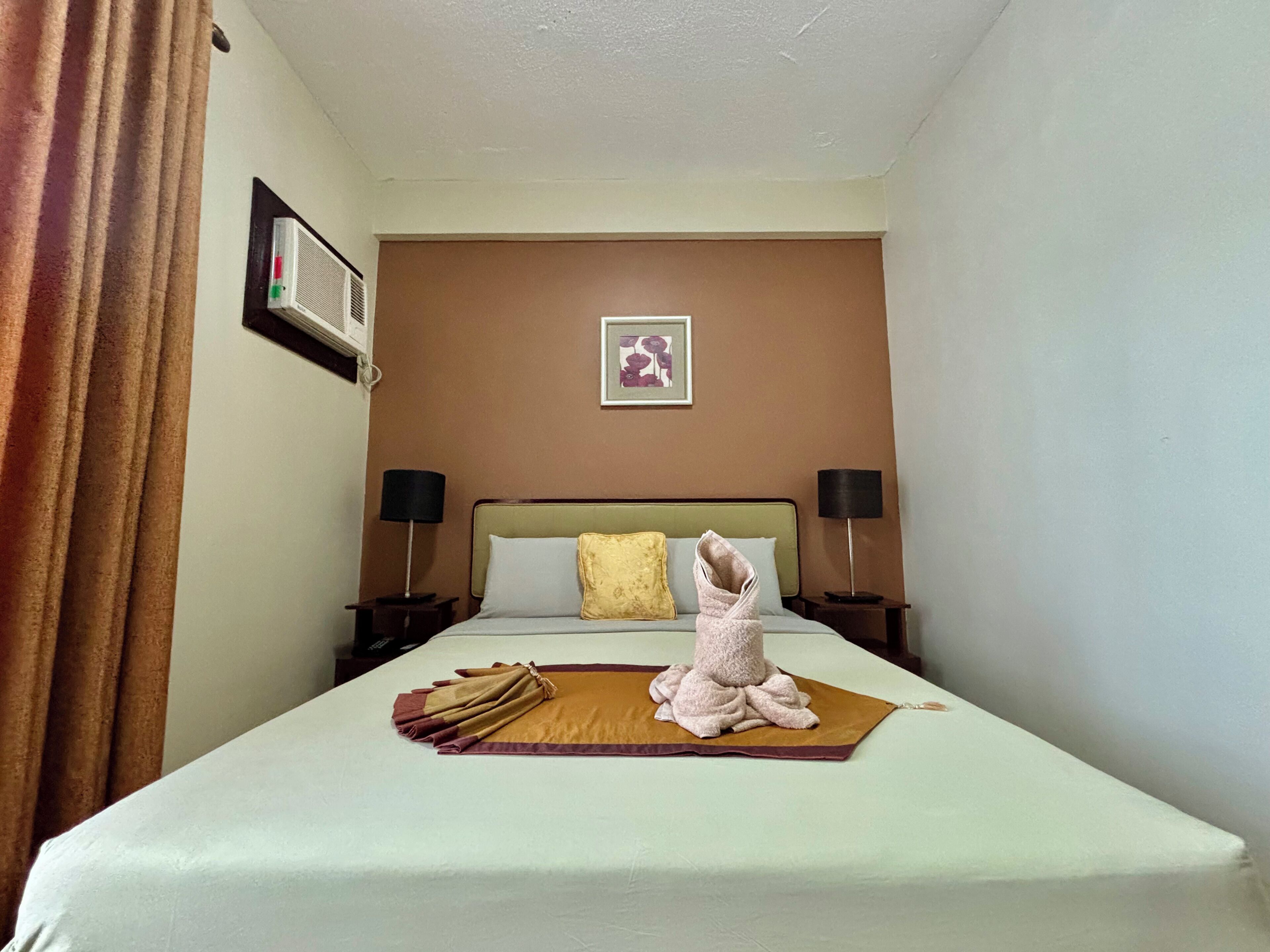 Une chambre d’hôtel avec un lit, deux tables de chevet, un climatiseur fixé au mur, un tableau encadré et un rideau.