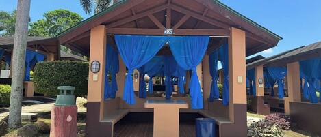 A covered outdoor seating area with blue curtains, a trash bin, and a bench.