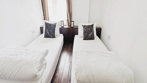 Habitación de hotel con dos camas individuales, ropa de cama blanca, suelo de madera oscura y una pequeña mesa con un florero entre las camas.