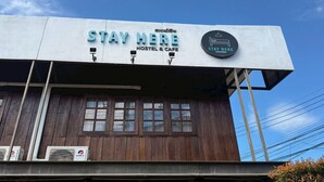 Un hostal y cafetería con un letrero que dice "STAY HERE HOSTEL & CAFE" y un logotipo circular con un icono de cama.
