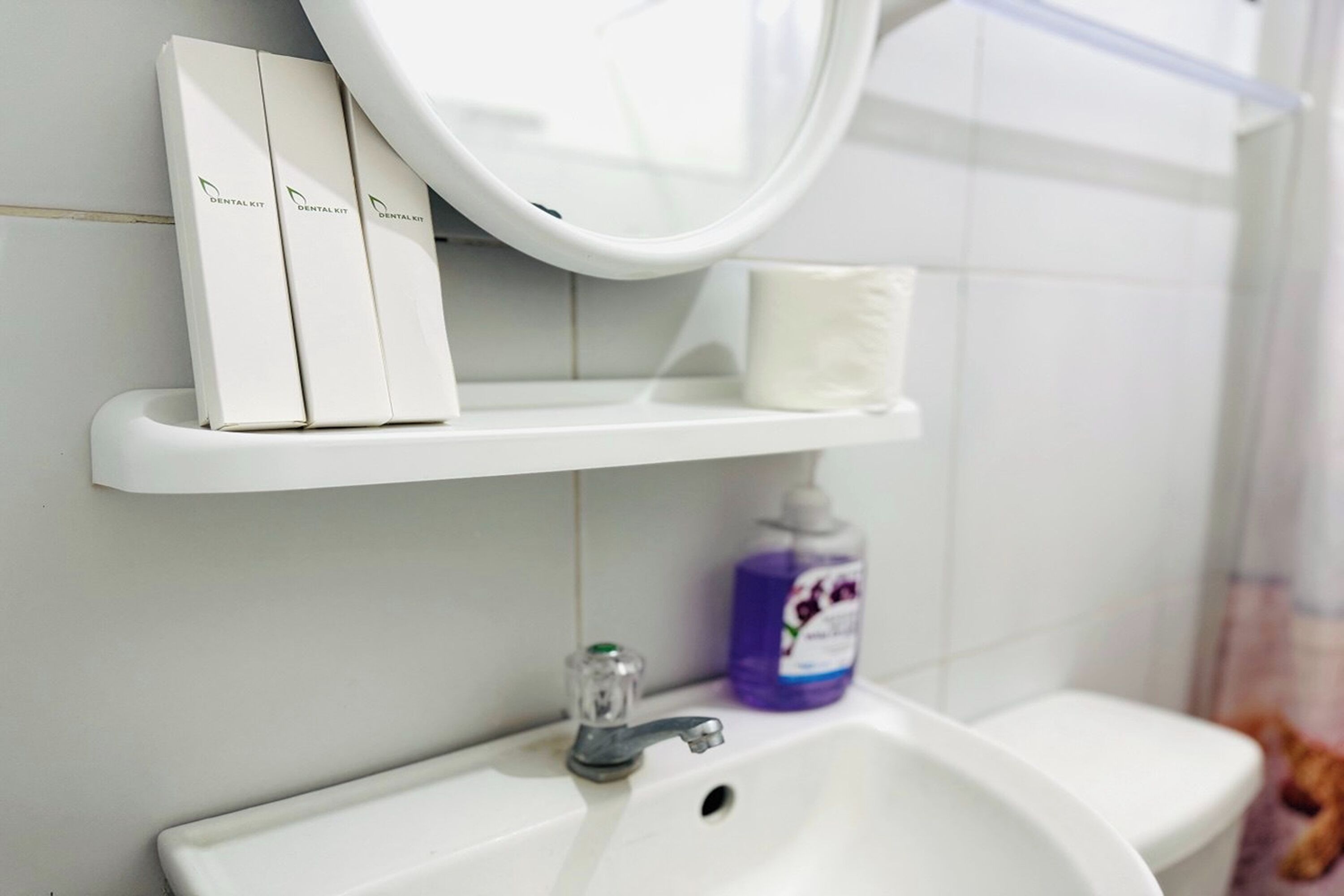 A bathroom with a white sink, a mirror, and a shelf holding dental kits and a bottle of hand soap.