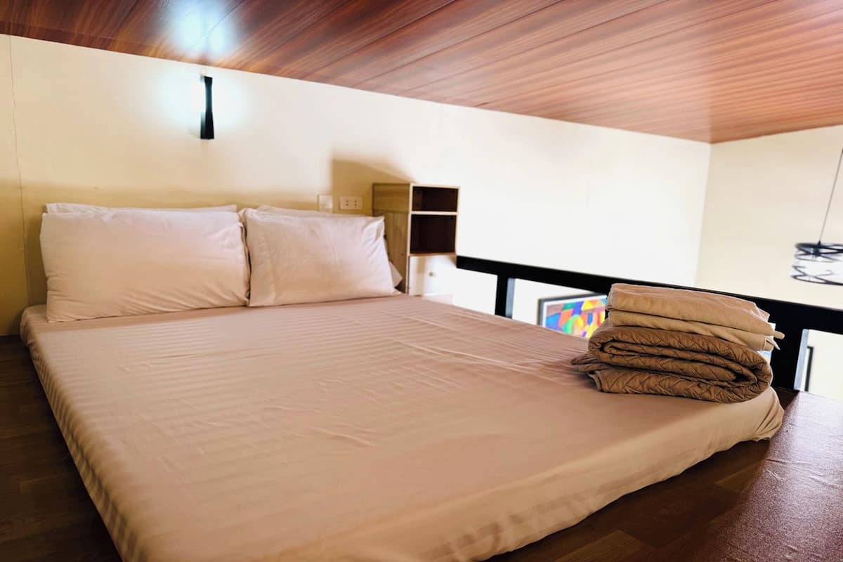 Una cama bien hecha con un edredón y almohadas de color claro, un cabecero de madera, una mesita de noche con una lámpara y una pila de mantas dobladas en el suelo.