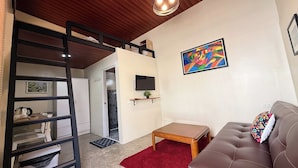 Un espacio habitable compacto con una cama individual superpuesta, una mesita pequeña, un sofá, una alfombra roja y un cuadro abstracto y colorido en la pared.