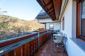 A balcony with a table and chairs, a view of a mountainous landscape, and a building with a tiled roof. - Apartment 'Himmelreich' with Shared Terrace, Garden and Wi-Fi (Oberharmersbach)