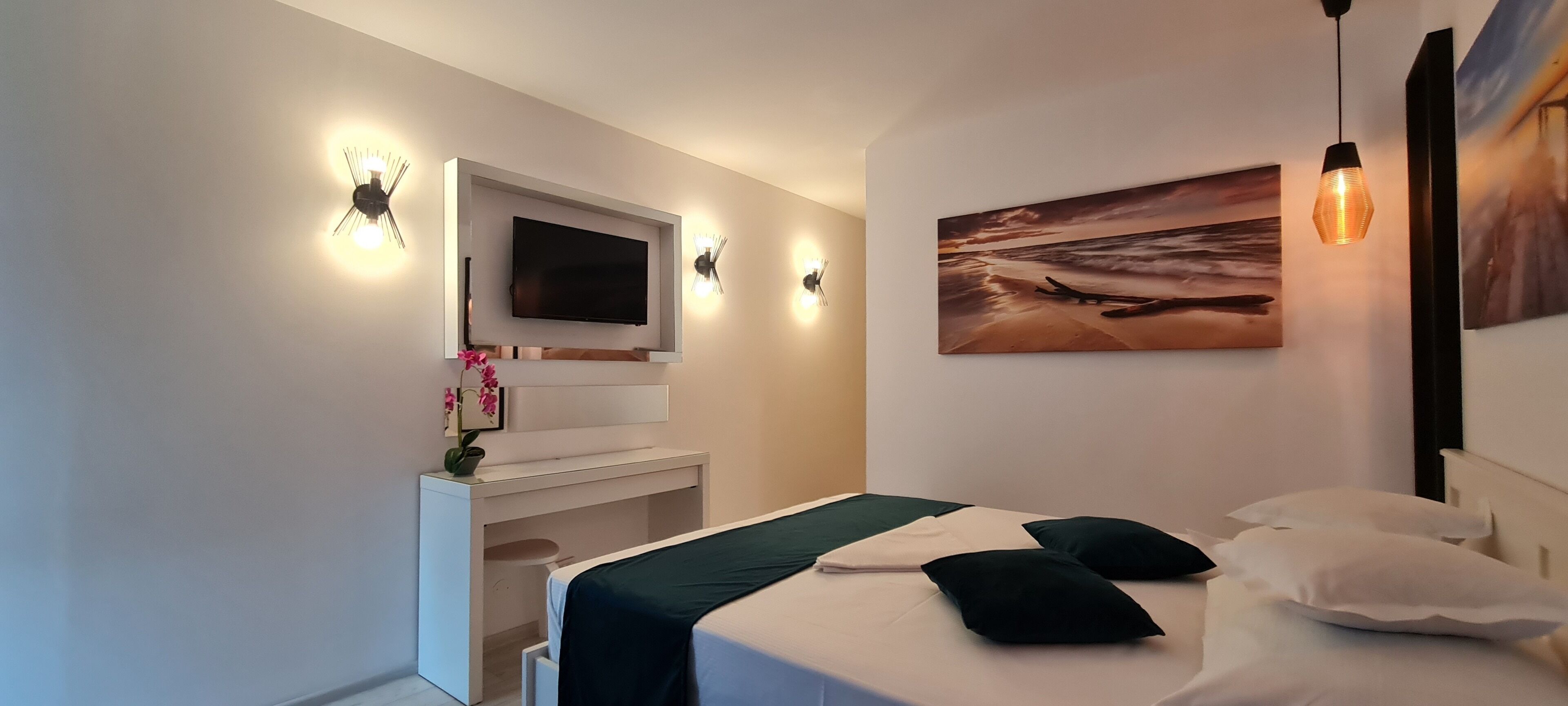 A modern hotel room with a flat-screen TV mounted above a white console, a bed with a green and white bedspread, two black pillows, and a painting of a sunset on the wall.
