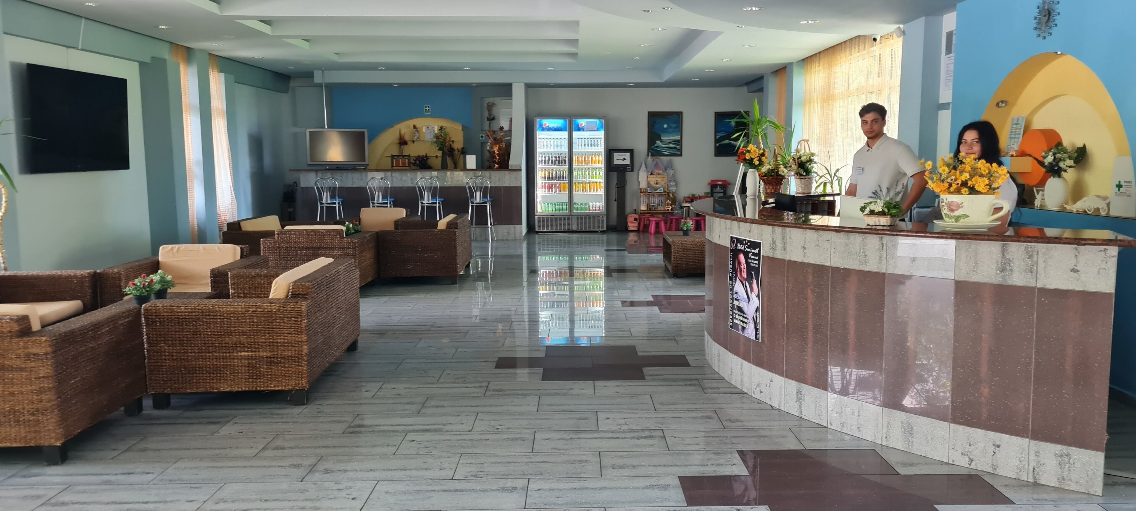 A hotel lobby with wicker furniture, a reception desk, a flat-screen TV, and a Pepsi refrigerator.