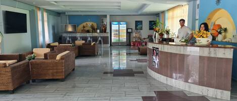 A hotel lobby with wicker furniture, a reception desk, a flat-screen TV, and a Pepsi refrigerator.