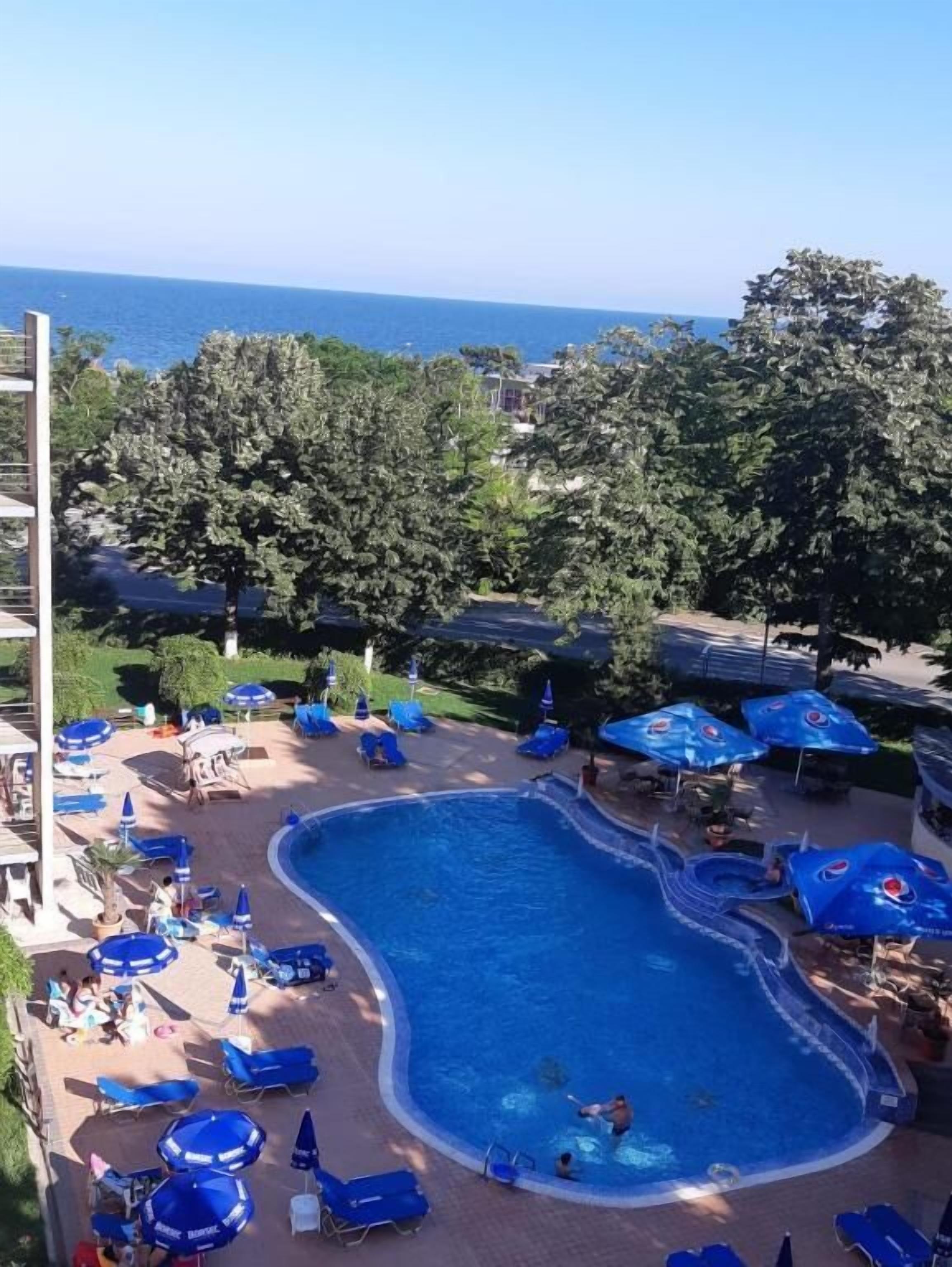 Ein Schwimmbecken mit Liegestühlen und Sonnenschirmen, umgeben von Bäumen und Blick aufs Meer.