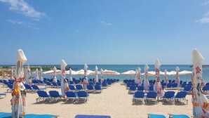 Ein Strand mit Reihen aus blauen Liegestühlen und weißen Sonnenschirmen mit bunten Mustern, auf einem sandigen Strand unter blauem Himmel.