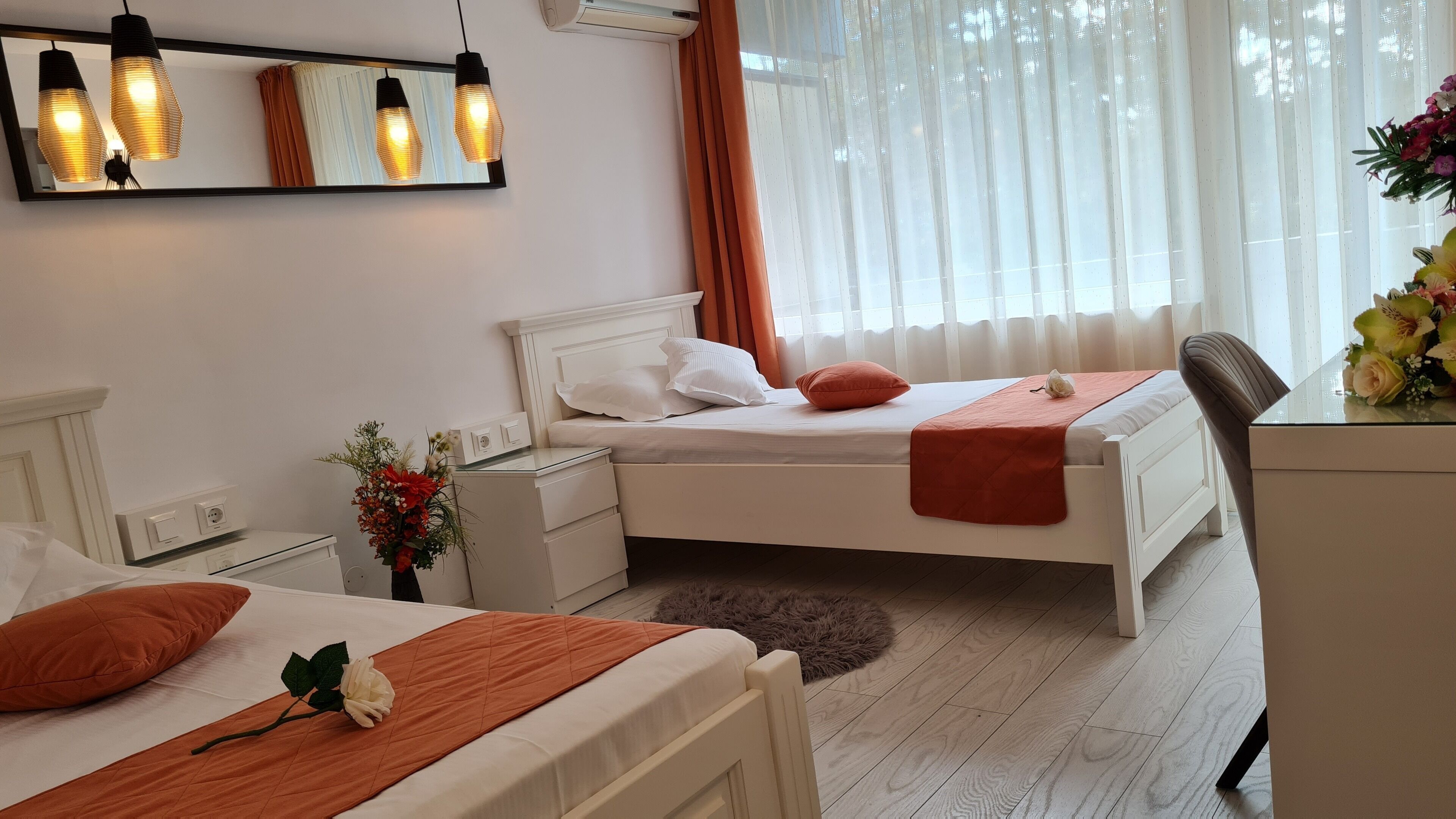 A bedroom with a bed, bedside tables, a desk, and a chair. There is a window with curtains, a mirror, and a wall-mounted light fixture.