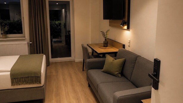 A hotel room with a bed, a sofa, a small table with a plant, a TV mounted on the wall, and a sliding door leading to a balcony. - UNO smart Soltau Wietzendorf (Wietzendorf)