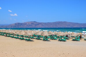 A beach with rows of green lounge chairs and thatched umbrellas, a clear blue sky, and a mountainous backdrop.