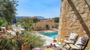 A poolside area with a stone wall, lounge chairs, a table with wine glasses, and a view of a mountain.