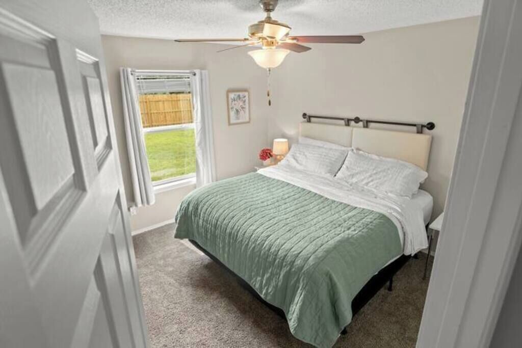 Ein Schlafzimmer mit einem Bett, einem Deckenventilator, einem Fenster mit Jalousien und einem Nachttisch mit Lampe.