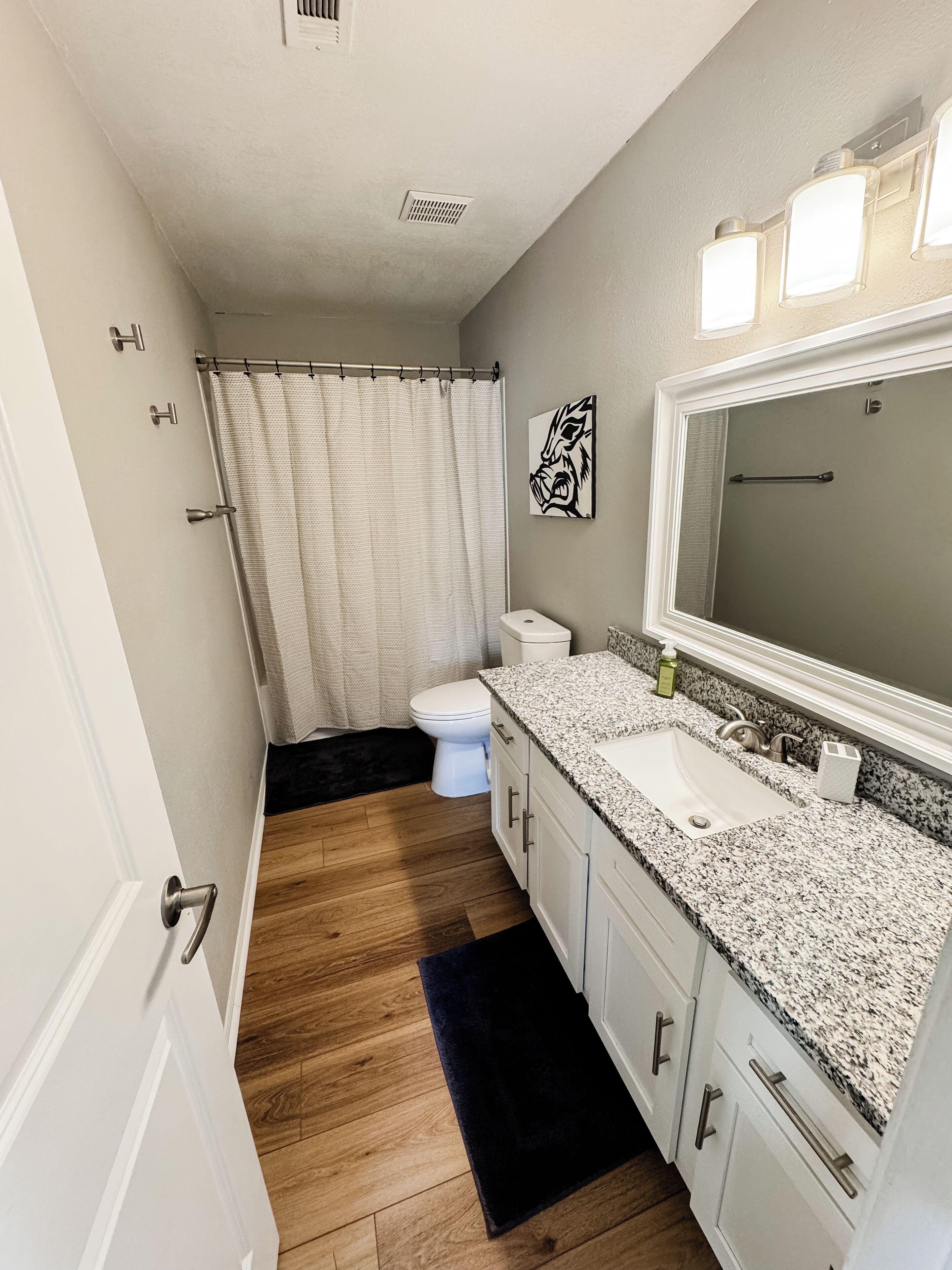 A bathroom with a white toilet, a shower with a white curtain, a granite countertop sink, and a large mirror.