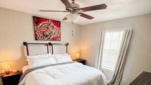 A bedroom with a bed, a nightstand, a ceiling fan, a window with curtains, and a wooden bench. - 3 KING BEDS Furr-Friendly Fayetteville Retreat (Fayetteville)