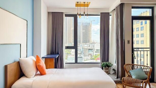 A modern hotel room with a large bed, a view of the cityscape, and a balcony with a wicker chair. - Sea Vista Boutique Hotel & Apartment By Haviland (Da Nang)