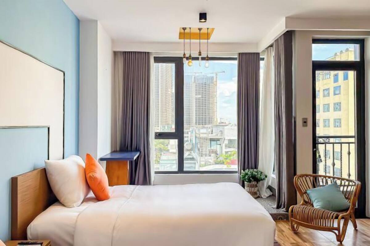 Quarto de hotel moderno com uma cama grande, vista para a paisagem urbana e uma varanda com cadeira de vime.