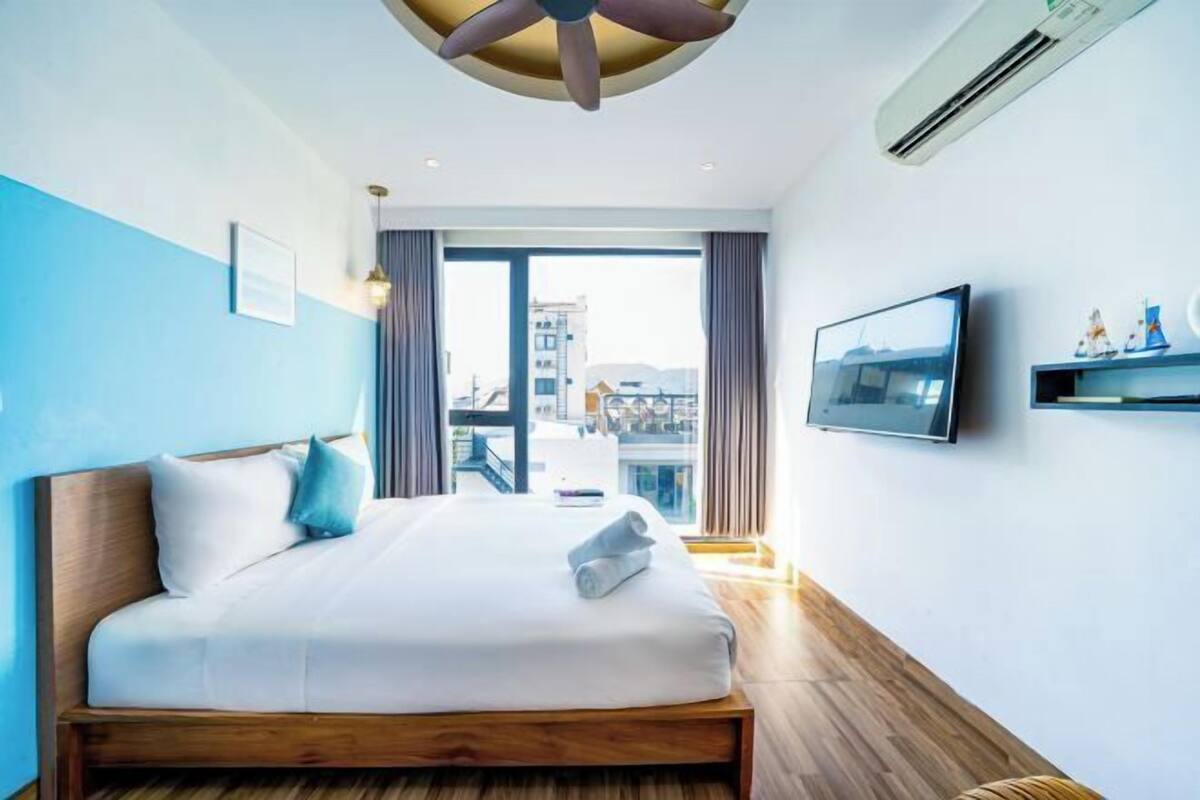 Quarto de hotel moderno com uma cama grande, ventilador de teto, ar condicionado, televisão e vista para a cidade.