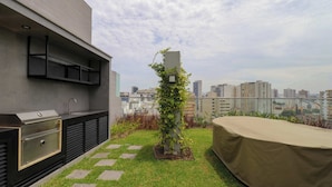 Eine Dachterrasse mit integriertem Grill, einem Pflanzgefäß mit Kletterpflanzen und Blick auf die Stadtlandschaft.