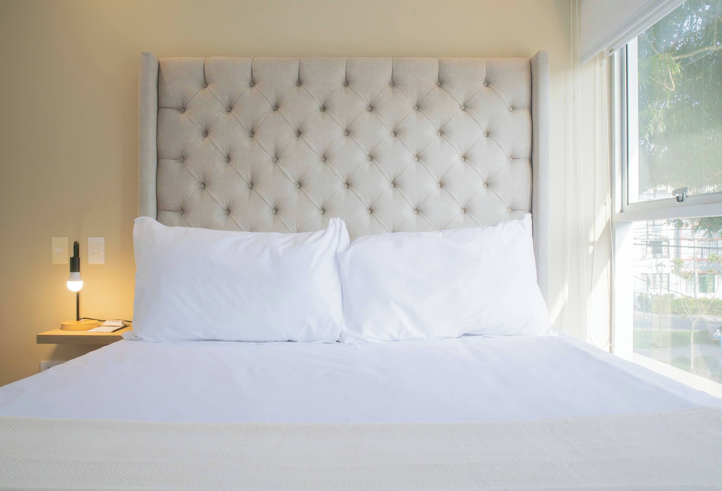 Una cama bien tendida con cabecera capitonada, ropa de cama blanca y una lámpara de noche.