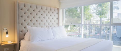 A neatly made bed with a tufted headboard, a bedside lamp, and a window with a view of trees and buildings.
